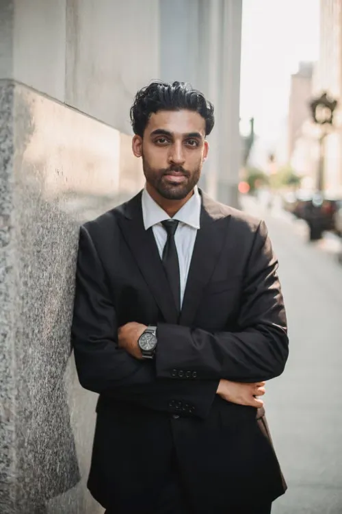 A man in a dark suit with arms crossed, leaning against a stone wall on a city street