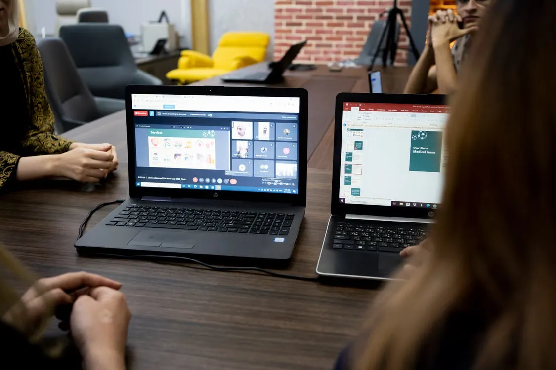 Two laptops on a meeting table, one showing a video call and one a presentation
