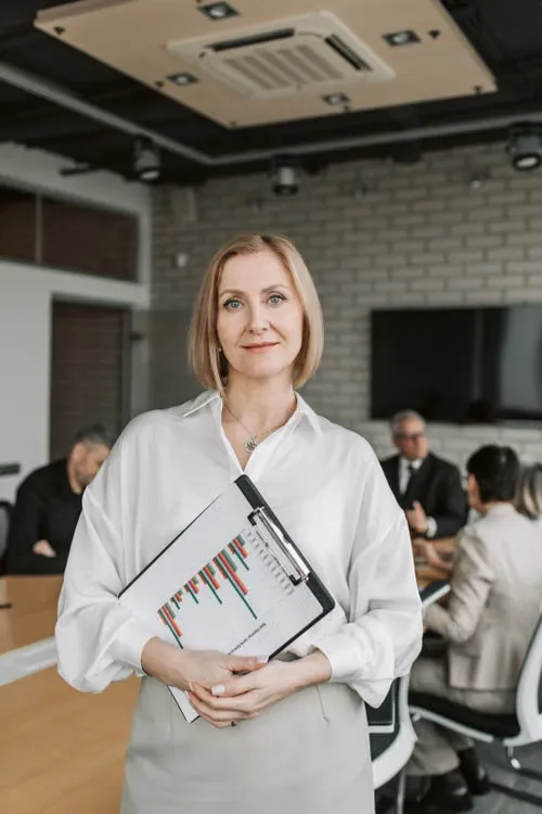 A businesswoman in a white shirt holding a clipboard with a bar chart, colleagues meeting behind her
